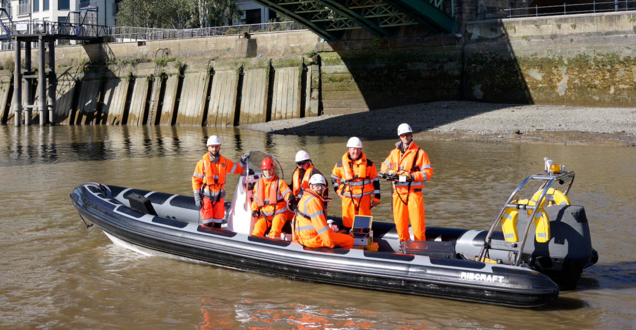 Survey and inspection team in high-visibility PPE operating from a Ribcraft workboat on a tidal river during marine engineering and infrastructure assessment.”