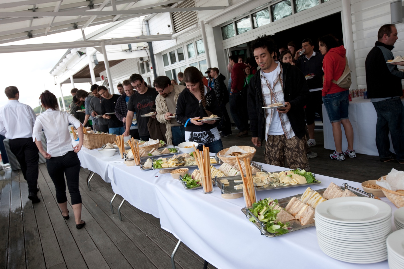 Guests line up at a long buffet table on an outdoor deck, choosing food from platters of sandwiches, salads, breadsticks and snacks, with a waterside venue and groups chatting in the background.