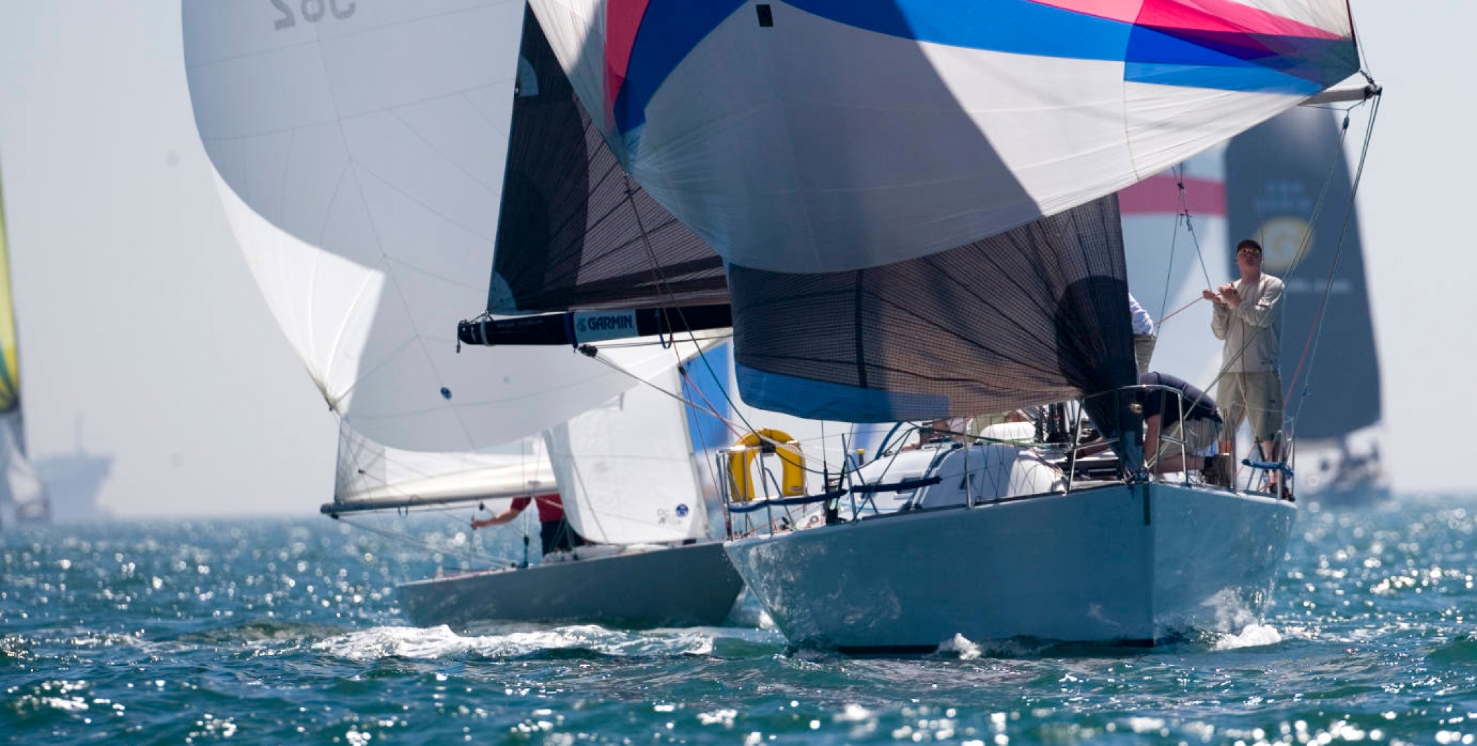 Performance yachts racing under colourful spinnakers on the Solent, with crew members working on deck during a sunny sailing event.