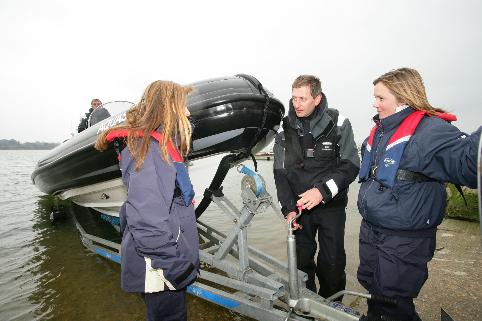 Instructor teaching two trainees how to safely launch a RIB from a boat trailer at the water’s edge, wearing waterproof gear and lifejackets.