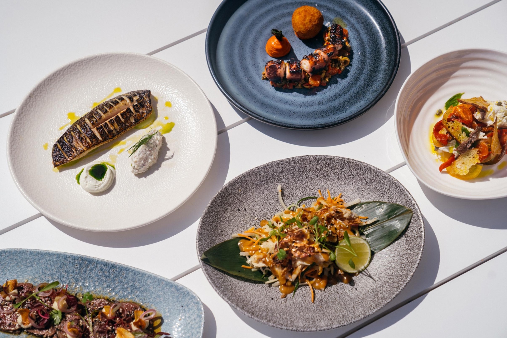 Selection of gourmet dishes at The Hut restaurant, featuring seafood, fresh salads, grilled fish and vibrant modern plating on a white outdoor table
