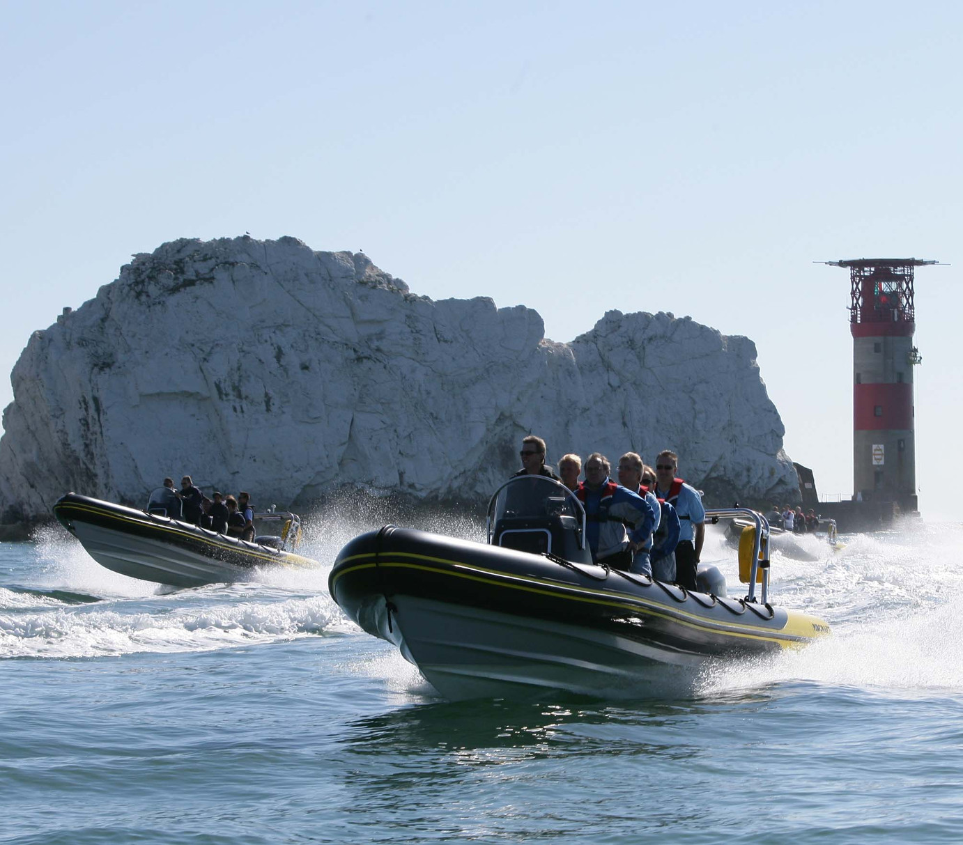 Two rigid inflatable boats (RIBs) filled with passengers speed across calm water near The Needles, the iconic white chalk cliffs. The red-and-white Needles lighthouse stands on the right, while sea spray rises around the boats as they travel in bright, clear weather.