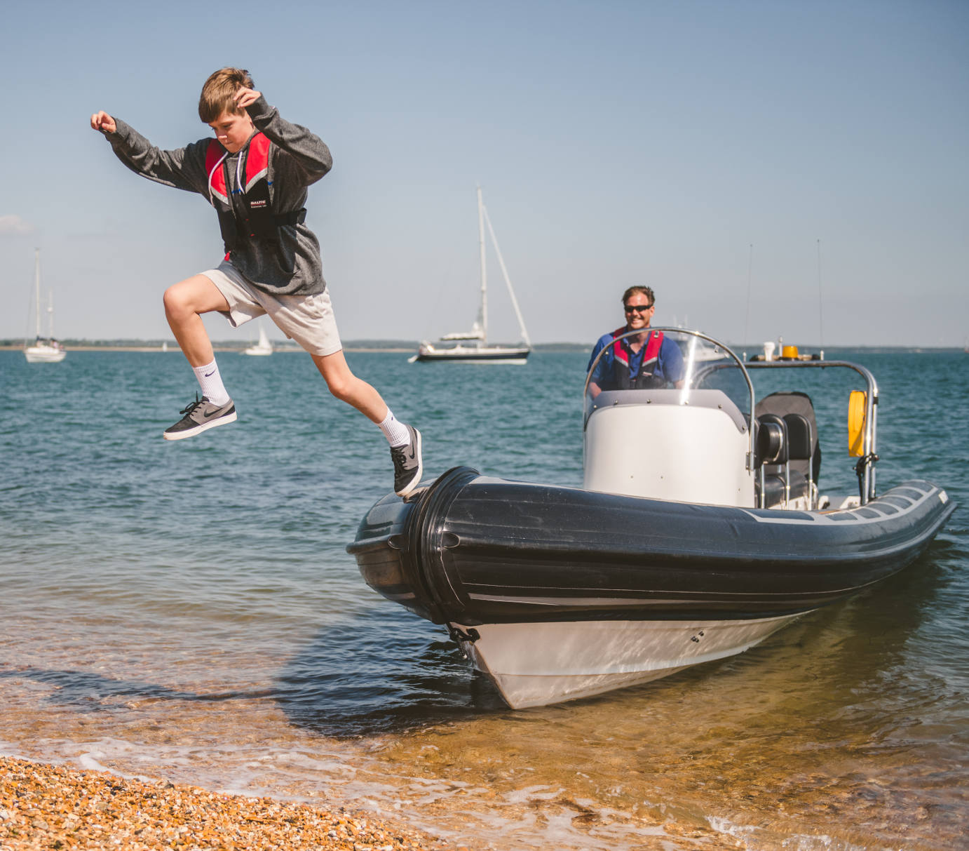 A boy wearing a life jacket leaps energetically from the bow of a rigid inflatable boat (RIB) onto a pebbled beach. The boat is gently beached at the shoreline, with an adult sitting at the helm behind him. Calm blue water and several anchored sailboats are visible in the background.