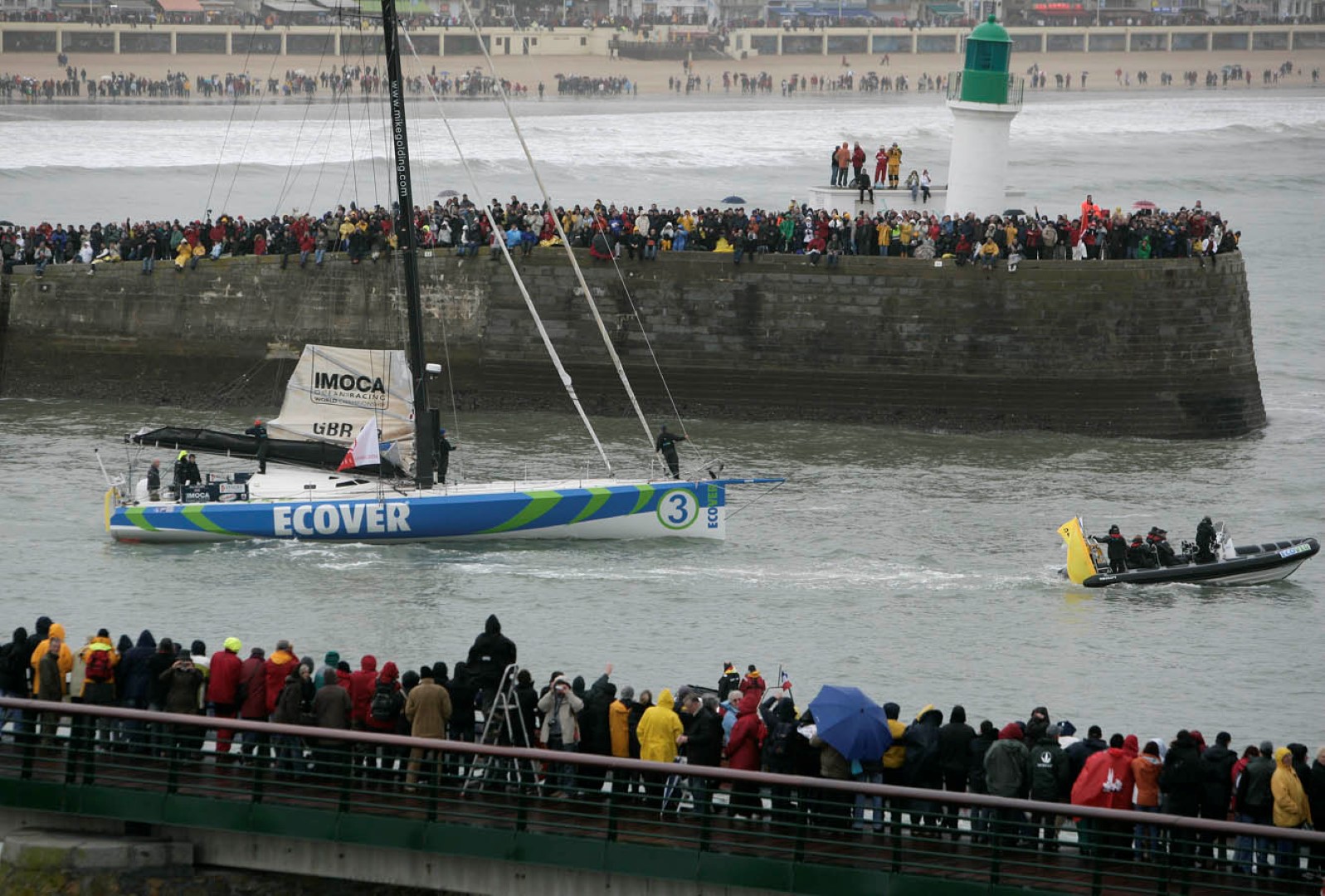 Large crowds gather along a harbour wall to watch an IMOCA racing yacht and a Solent Rib Charter support RIB navigate out to sea during a major sailing event.