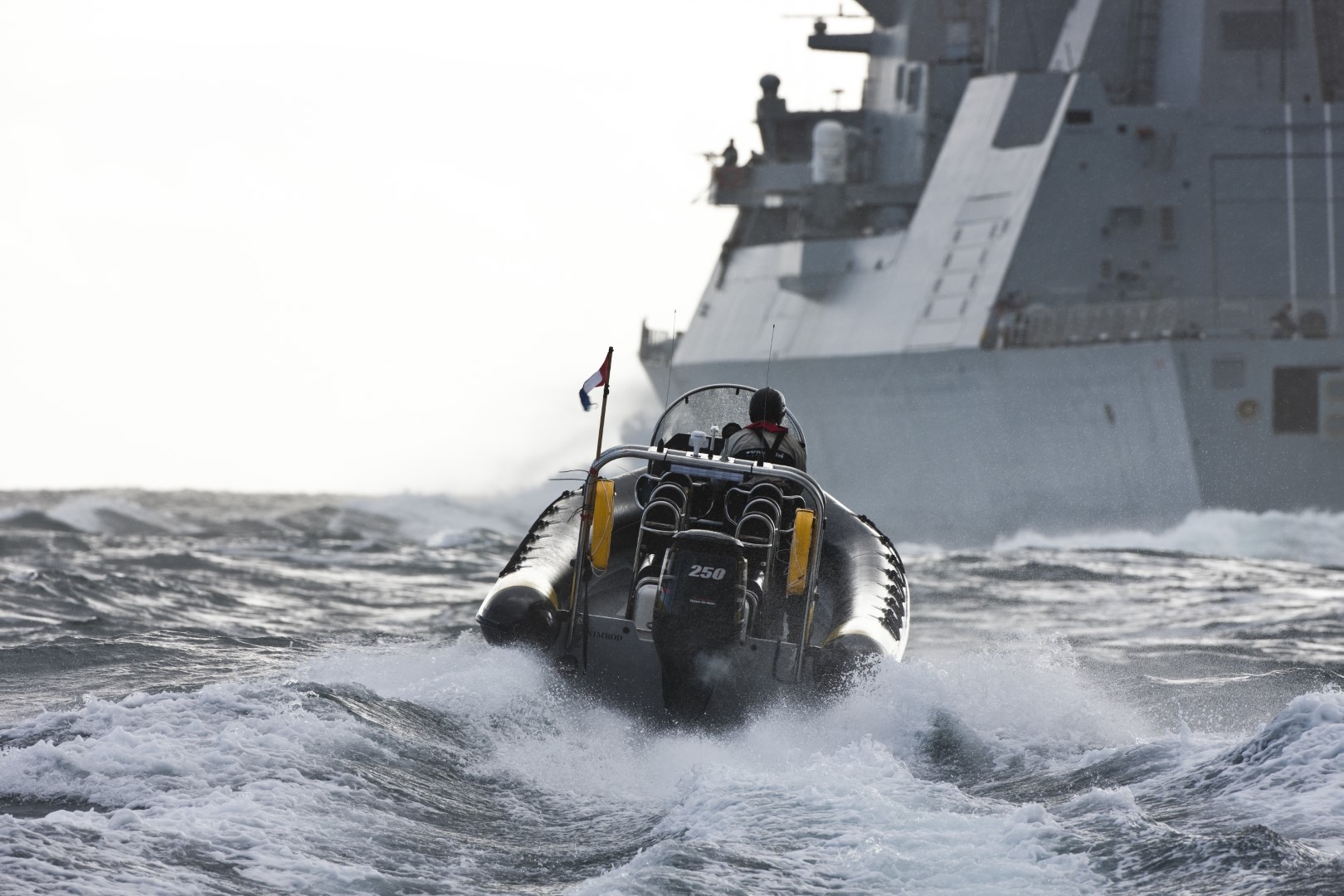 Rigid inflatable boat travelling through rough seas with a naval vessel in the background