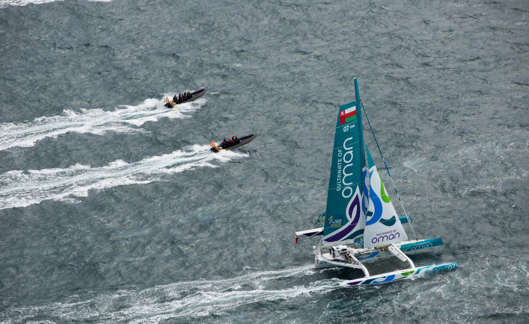 A group of passengers in waterproof gear waving and smiling aboard a Solent Rib Charter RIB as it speeds across the Solent with sailing boats in the background.