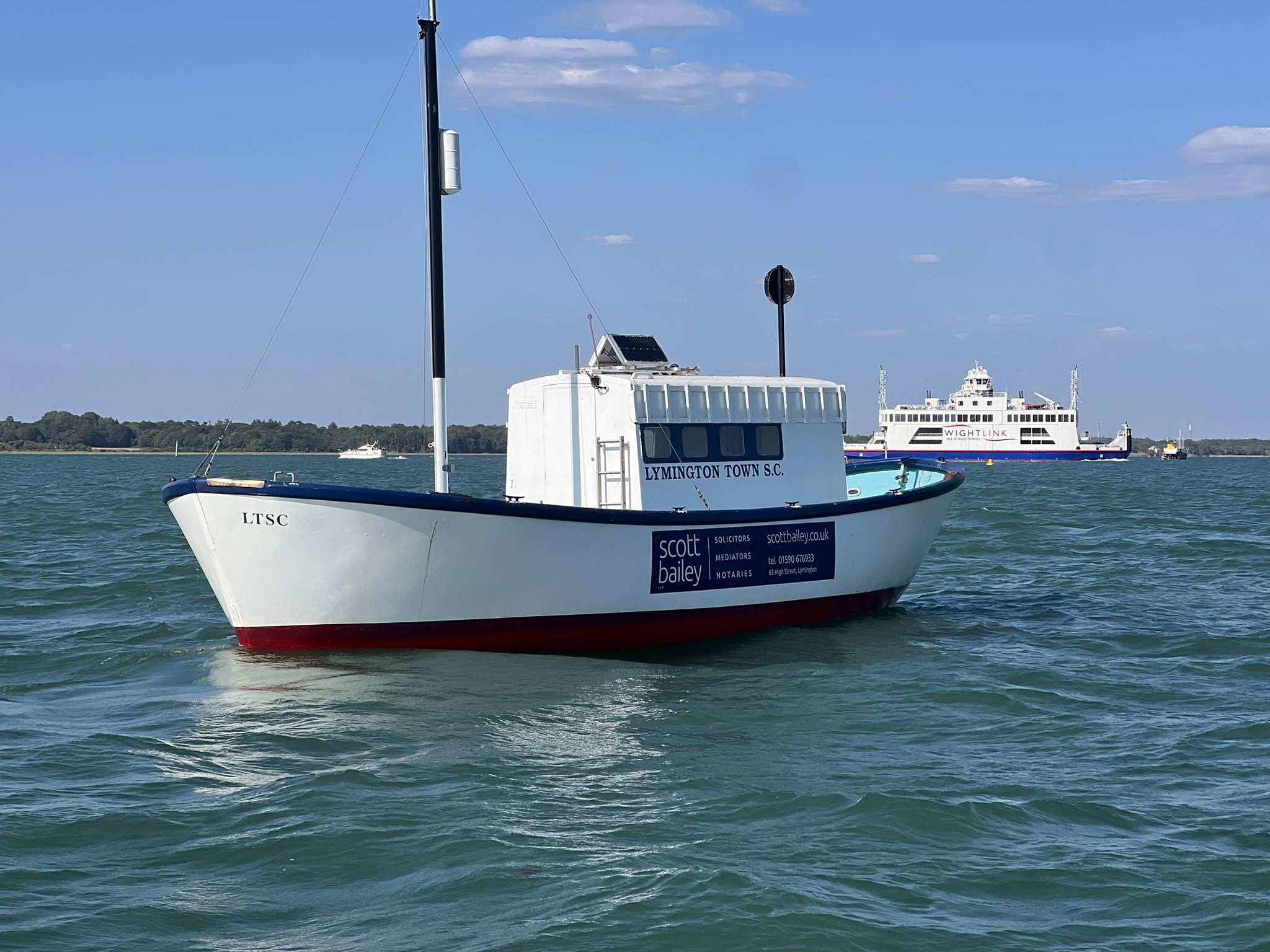 The Baverstock, a small white-and-blue boat marked “LTSC” and “Lymington Town S.C.,” floats on choppy blue-green water, with a Wightlink ferry in the background beneath a bright blue sky.